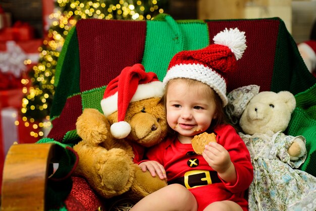 Rosto de bebê fofo com chapéu de Papai Noel, perto de árvore de Natal. Crianças de ano novo. Conceito de infância, infância e pessoas - bebê feliz com chapéu de Papai Noel sobre fundo de luzes de férias.