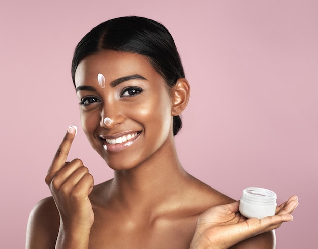 Rosto de cuidados com a pele e mulher com recipiente de creme no estúdio isolado em um fundo rosa Creme de dermatologia de produto e retrato de modelo feminino indiano feliz aplicam loção hidratante e cosméticos