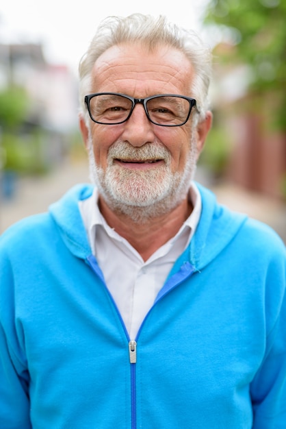 Foto rosto de homem barbudo sênior bonito feliz sorrindo enquanto usava jaqueta azul e óculos ao ar livre