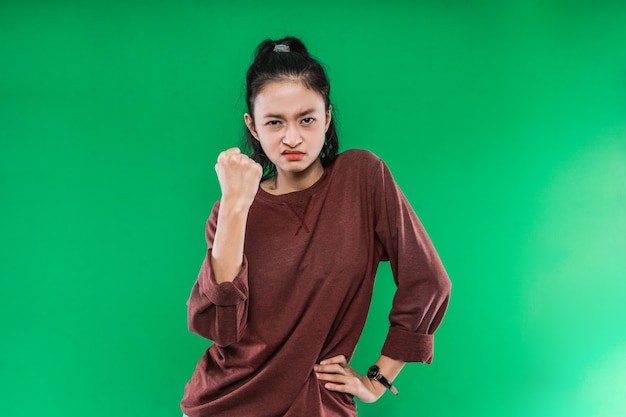 Rosto de mulher jovem com uma expressão de raiva enquanto fecha um punho e uma mão na cintura contra um fundo verde