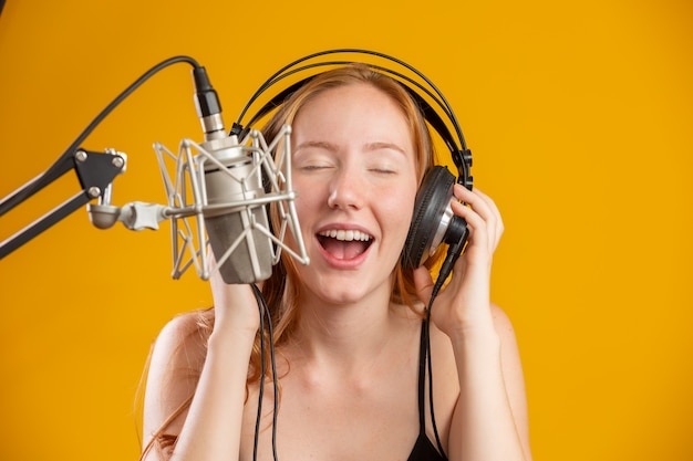 Rosto de mulher ruiva linda cantando com uma boca aberta do microfone prata condensador realizando pose de música sobre espaço de cópia de parede amarela para o seu texto. locutor de rádio fm.