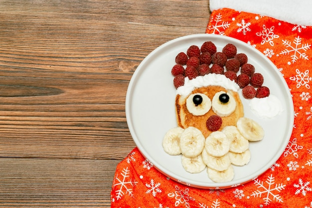 Rosto de Papai Noel de Natal em forma de panqueca com doce de framboesa fresca, banana no prato, meia de Natal em fundo de madeira para o café da manhã de crianças de crianças. sobremesa comida de natal, decorações de ano novo