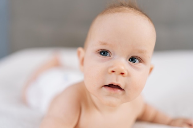 Rosto em close-up de um bebé atraente de fraldas brancas deitado de barriga felizmente agita os braços e