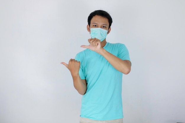 Rosto feliz de jovem asiático usa máscara médica e azul com ponto de mão no espaço vazio