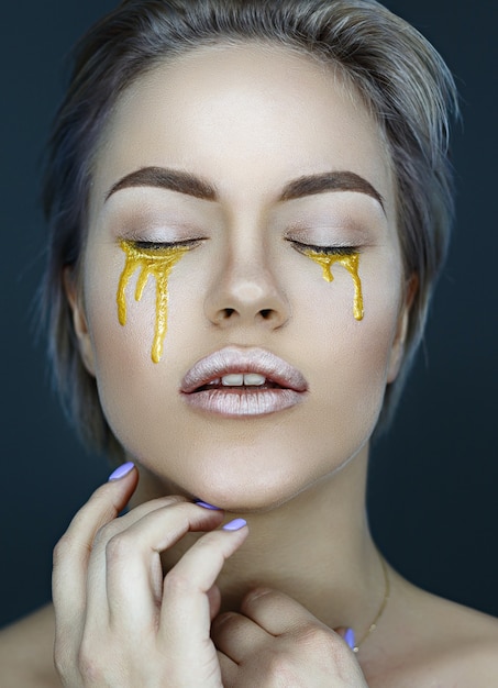 Rosto feminino jovem com maquiagem artística dourada, olhando para a câmera, vista frontal do close up das lágrimas