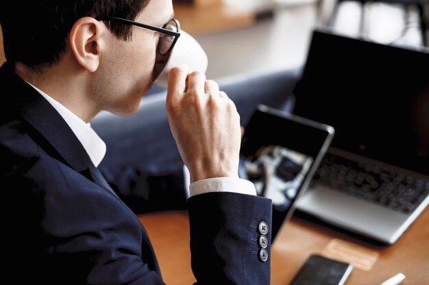 Rosto masculino, bebendo café, segurando um tablet sentado em uma mesa com o laptop nele.