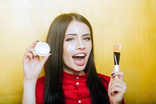 Rosto sorridente de mulher sexy e fofa ou menina bonita com cabelo comprido morena e batom nos lábios tem maquiagem da moda em fundo amarelo, possui escova de artista e frasco de creme