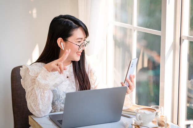 Rosto sorridente feliz asiáticos freelance pessoas Mulher de negócios usando fones de ouvido sem fio usando a câmera de videochamada do smartphone e conversando enquanto trabalha casualmente com o computador laptop no café, Estilo de vida