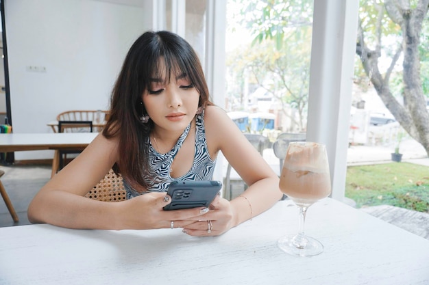 Rosto triste de uma jovem asiática segurando um telefone celular vestindo uma blusa sentada perto de uma janela Conceito de propaganda