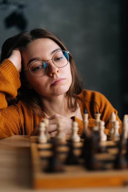 Rosto vertical fechado de uma jovem pensativa em óculos elegantes pensando em movimento de xadrez sentado no foco seletivo do quarto escuro