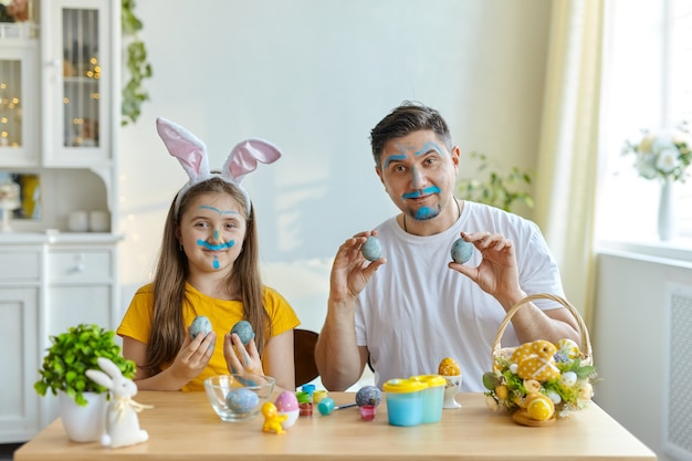 Rostos de pai e filha manchados de tinta azul para pintar ovos. sobre a mesa está uma cesta com ovos de Páscoa e tintas.