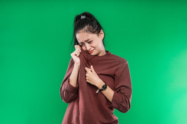 El rostro de una expresión de llanto de una joven asiática mientras se limpia las lágrimas con ropa aislada sobre fondo verde