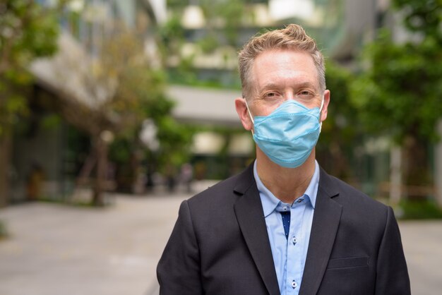 Rostro de hombre de negocios maduro con máscara para protegerse del brote de coronavirus con la naturaleza en la ciudad