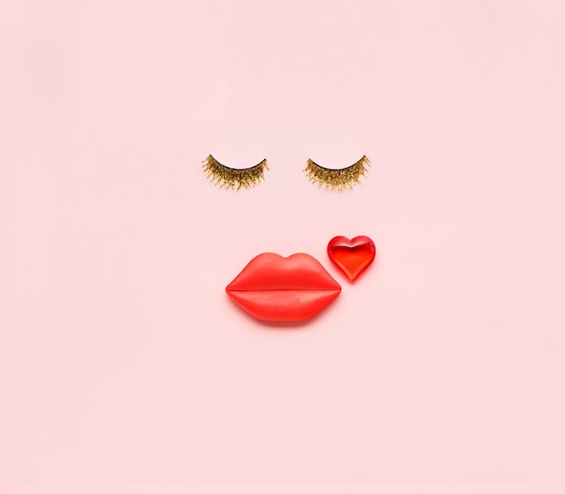 Rostro de mujer divertida creativa hecha de pestañas brillantes, corazones y labios rojos sobre un fondo rosa. Concepto de belleza mínima, tarjeta del día de San Valentín. Vista plana endecha, superior.