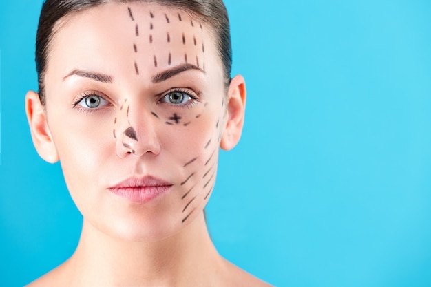 Rostro de mujer hermosa. retrato de belleza de mujer joven morena sonriente, líneas pintadas en la cara para cirugía estética sobre fondo azul. cirujano estético blefaroplastia. rinoplastia