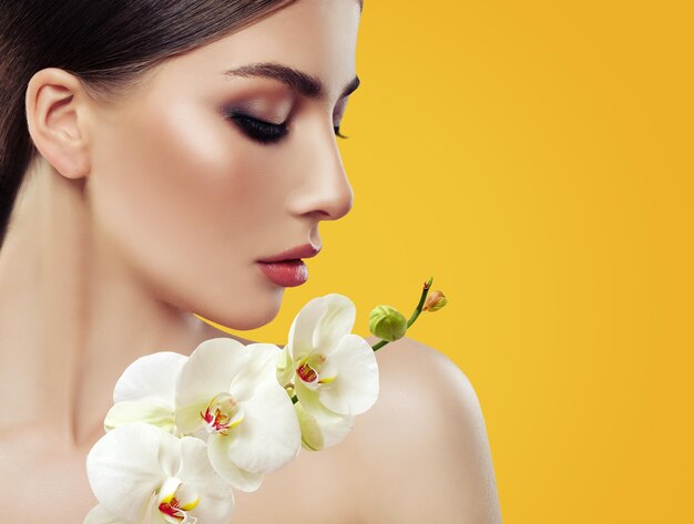 Rostro de mujer joven perfecta con maquillaje y flores de orquídeas sobre fondo amarillo brillante Retrato de belleza