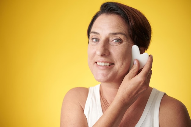 Rostro de mujer madura feliz haciendo masaje con herramienta gua sha