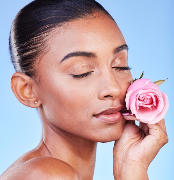 Rostro de rosa y cuidado de la piel de una mujer en un estudio para cosmética ecológica, bienestar natural o fondo azul. Modelo indio con flores y ojos cerrados para la belleza vegana. Dermatología sostenible y estética floral.