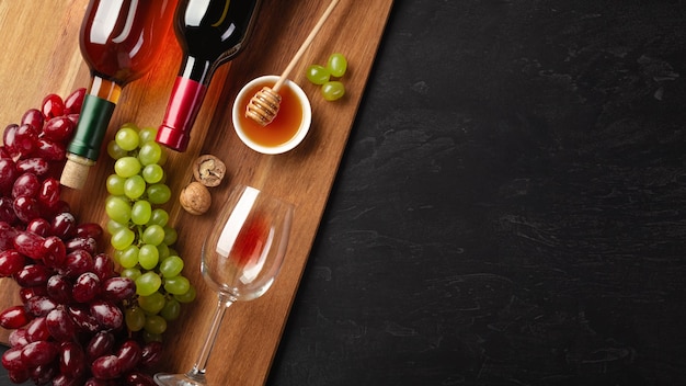 Rot- und Weißweinflaschen mit Trauben, Käse, Honig, Nüssen und Weinglas auf Holzbrett und schwarzem Hintergrund. Draufsicht mit Kopienraum.