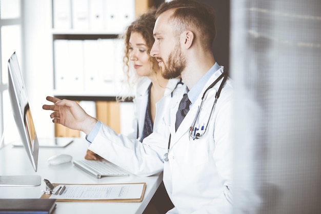 Rotbärtiger Arzt mit Kollegin bespricht aktuelle Krankheitstherapie, während er am Arbeitsplatz in der Klinik sitzt. Teamarbeit in der Medizin.