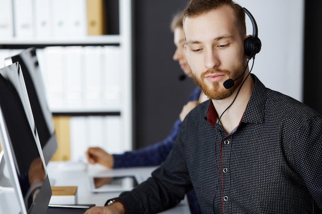 Rotbärtiger Geschäftsmann spricht per Headset in der Nähe seiner Kollegin, während er in einem modernen Büro sitzt. Verschiedene Personengruppen im Call Center. Telemarketing und Kundenservice.