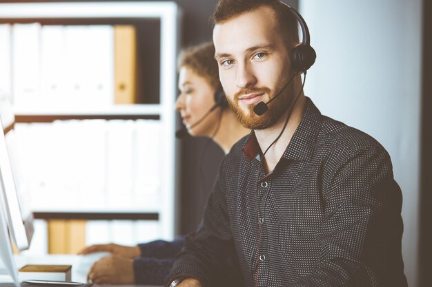 Foto rotbärtiger geschäftsmann spricht über headset neben seiner kollegin, während er im sonnigen büro sitzt. gruppe verschiedener menschen im callcenter.