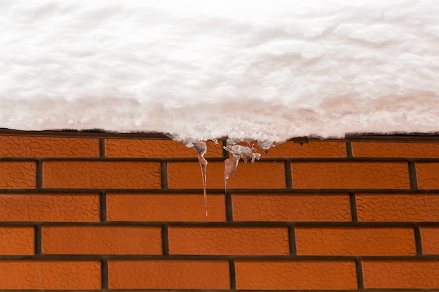 Rote Backsteinmauer. Textur. Die Dachrinne auf dem Schneedach. Eiszapfen hängen vom schneebedeckten Dach. Gefahr hängt vom Dach.