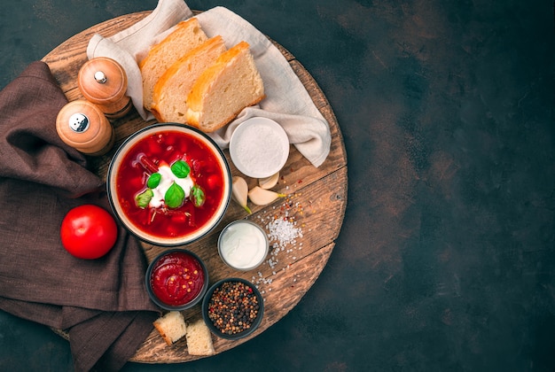 Rote-Bete-Suppe mit saurer Sahne und Basilikum, Brot, Tomate und Tomatensauce auf einem runden Holzbrett auf einem braunen Betonhintergrund. Seitenansicht.