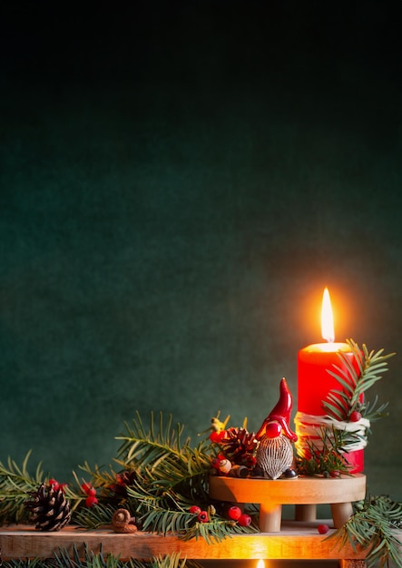 Foto rote brennende kerze mit keramikzwerg in rotem hut mit weihnachtszweigen auf dunkelgrünem hintergrund