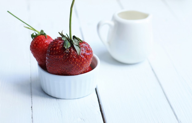 Rote Erdbeeren in der keramischen weißen Platte und im weißen Milchkrug für Creme auf weißem hölzernem Hintergrund.