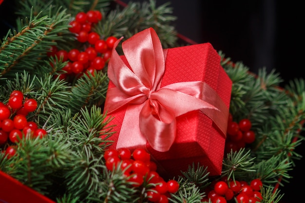 Rote Geschenkbox mit Schleife in Satinrosa, eingetaucht in die Nadeln eines Weihnachtsbaumes, verziert mit roten Beeren.