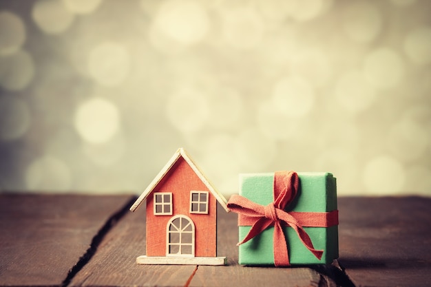 Rote Geschenkbox und Spielzeughaus auf Holztisch mit Bokeh im Hintergrund.