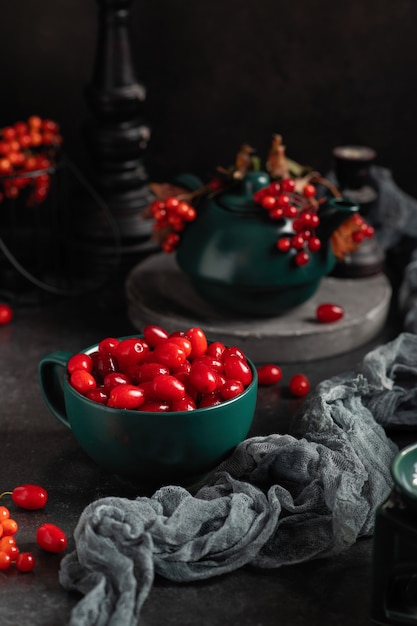 Foto rote hartriegelbeeren in einer grünen tasse mit einer teekanne, auf grauem hintergrund, hochformat