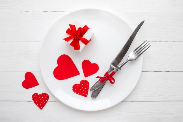 Rote Herzen auf einem weißen Teller, einer Gabel, einem Messer, einem roten Band, einer Geschenkbox auf einem weißen Holztisch