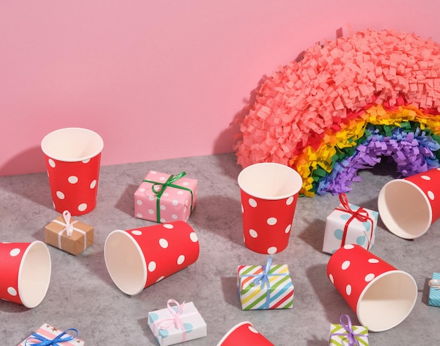 Rote Papierbecher mit weißen Punkten und auf dem Tisch verstreuten Geschenken. Bunter Pinata-Regenbogen für eine festliche Stimmung