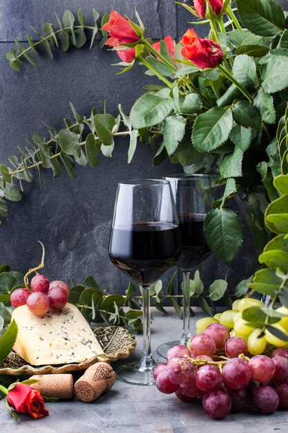 rote Rosen, Trauben, Käse und ein Glas Rotwein auf einem schwarzen konkreten Hintergrund