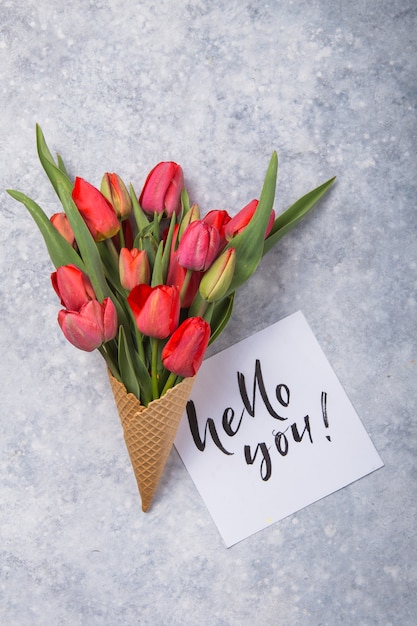 Rote schöne Tulpen in einer Eiscreme-Waffeltüte mit Karte Hallo Sie auf einem konkreten Hintergrund. Konzeptionelle Idee eines Blumengeschenks. Frühlingsstimmung