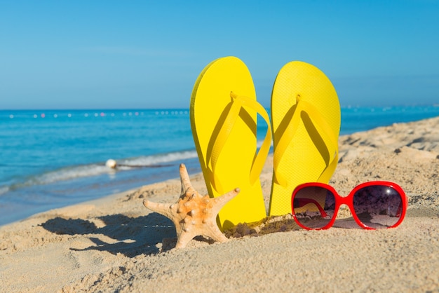 Rote Sonnenbrillen für Damen und gelbe Flip-Flops am Sandstrand. Reisen Sie auf dem Seeweg. Strandurlaub