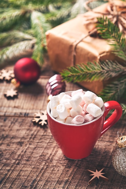 Rote Tasse heiße Schokolade mit Marshmallow- und Tannenzweigen, Geschenkboxen auf verschneitem Winterhintergrund mit schneebedeckten Zweigen auf altem Holztisch. Weihnachts- oder Winterkonzept.