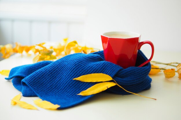 Rote Tasse mit Kaffee auf dem Tisch in einem blauen Strickschal und gelben Herbstblättern. Hallo Herbstkonzept. saisonale Krankheiten