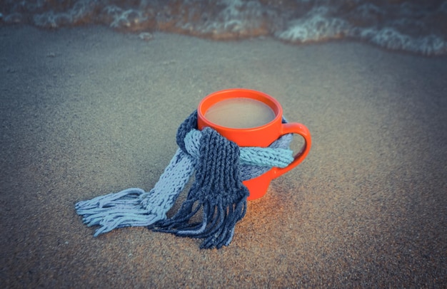 Rote Tasse mit Kaffee mit blauem Strickschal gebunden steht am Sandstrand