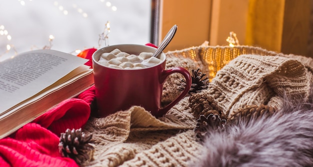 Rote Tasse mit Kakao und Marshmallows auf einem Holztisch mit Schals altes Buch nach Hause Komfort