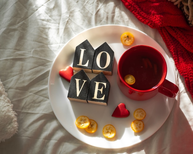 Rote Tasse Tee mit Kumquat auf dem Teller und zwei Herzplätzchen mit dem Wort LIEBE auf einem weißen Bett