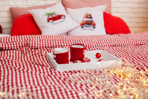Rote Tassen, die auf Holztablett mit Weihnachtsgirlande bleiben, liegen auf Bett