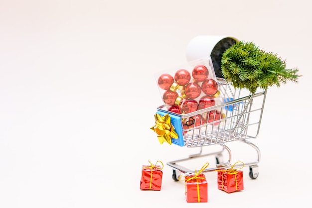 Rote Weihnachtskugeln und ein künstlicher kleiner Weihnachtsbaum mit Geschenken im Einkaufswagen auf einem hellen Pi...