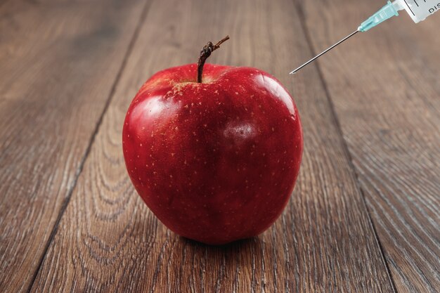 Roter Apfel, der eine Nadel oder eine Spritze und chemische Schädlingsbekämpfungsmittel auf einem hölzernen Hintergrund einspritzt