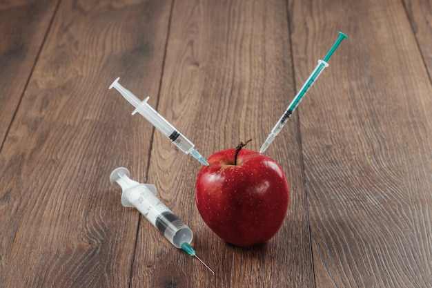 Roter Apfel, der eine Nadel oder eine Spritze und chemische Schädlingsbekämpfungsmittel auf einem hölzernen Hintergrund einspritzt