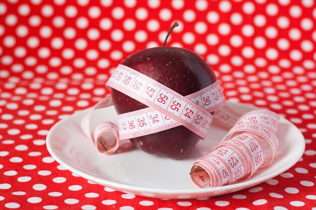 Roter Apfel, eingewickelt in ein Maßband, das auf einem weißen Teller auf einem roten Tupfen liegt