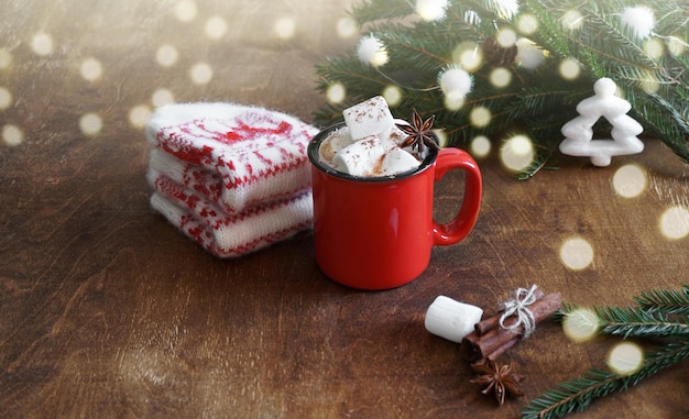 Roter Becher mit heißer Schokolade mit Marshmallow und Handschuhen und Zweigen von Weihnachtsbaum und Bokeh auf Holztisch