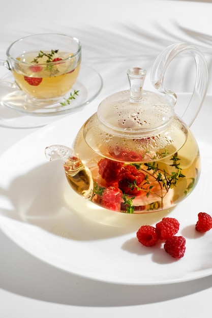 Roter Beerenkräutertee mit Thymian und frischer Himbeere in Teekanne aus Glas. Weißer Hintergrund mit Exemplar